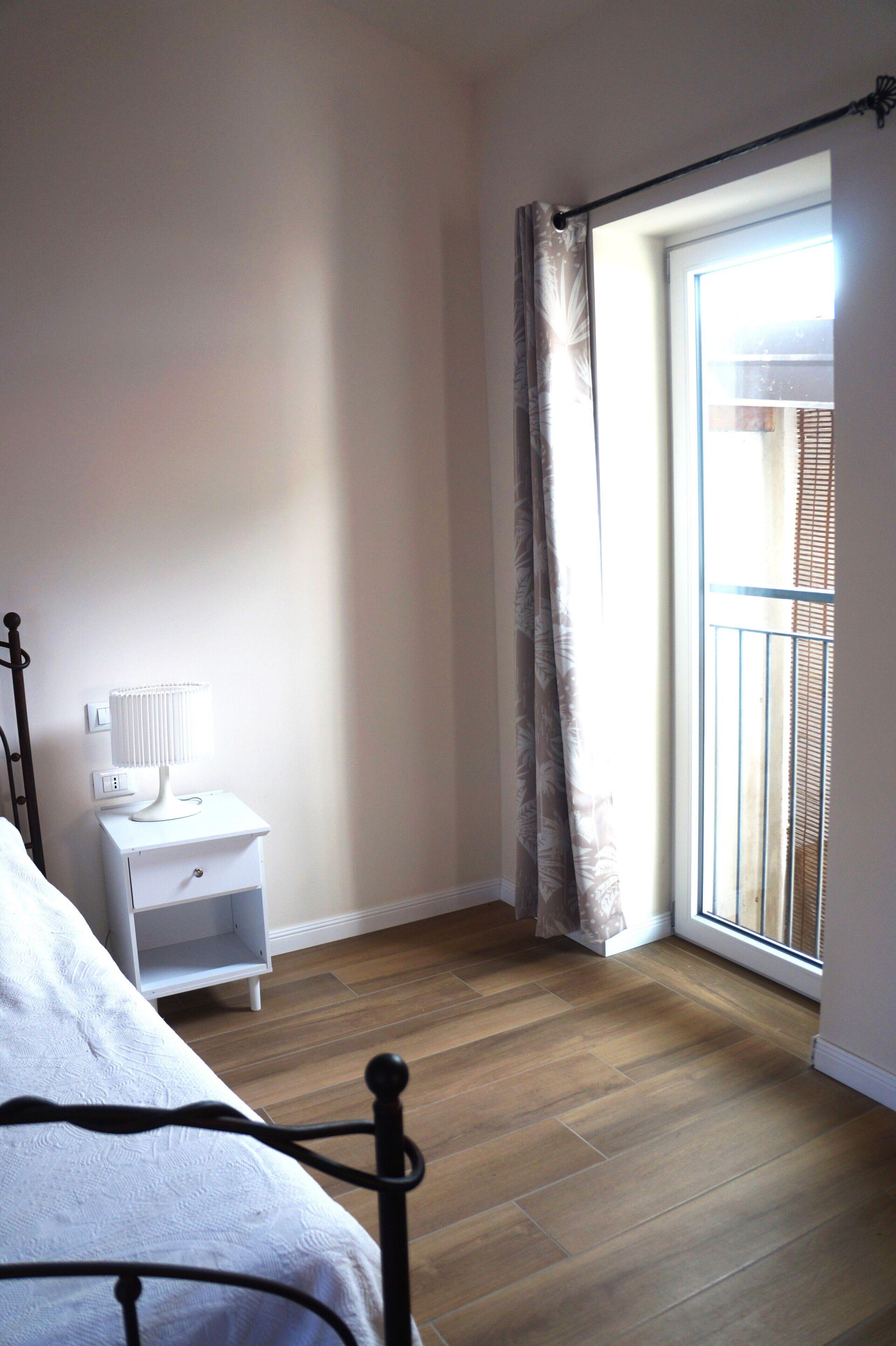 A bedroom with a bed, a bedside table with a lamp, a window with curtains, and a view of an outdoor balcony.