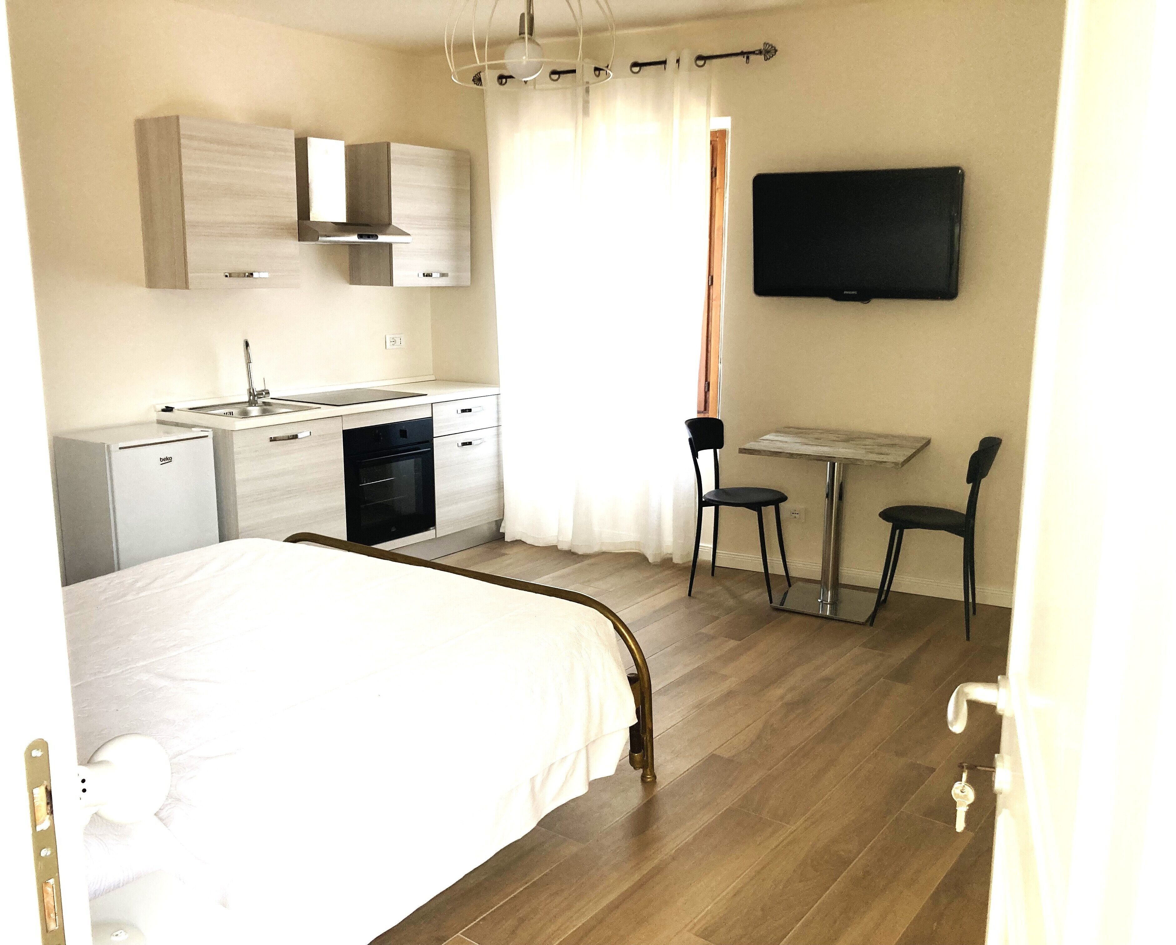 Habitación de hotel con cama, kitchenette, mesa de comedor con sillas, televisor y ventana con cortinas.