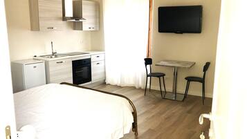 Habitación de hotel con cama, kitchenette, mesa de comedor con sillas, televisor y ventana con cortinas.