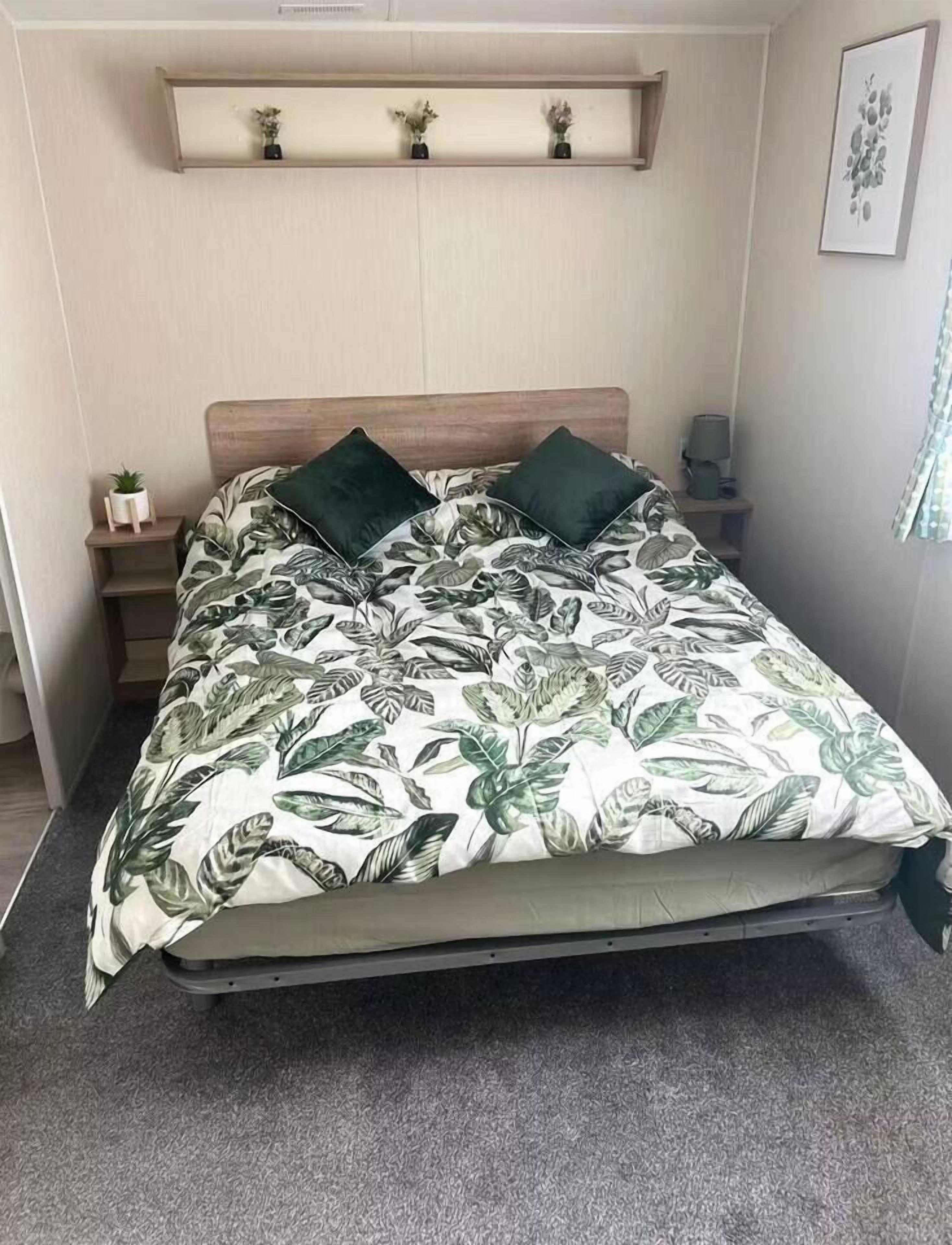 A bed with a leaf-patterned bedspread, a wooden headboard, a bedside table with a lamp, and a shelf above the bed.