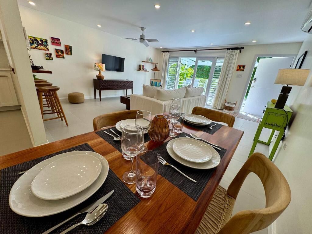A dining area with a wooden table set for four, a sofa, a TV, and a view of greenery outside.