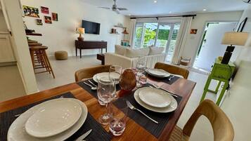 A dining area with a wooden table set for four, a sofa, a TV, and a view of greenery outside.