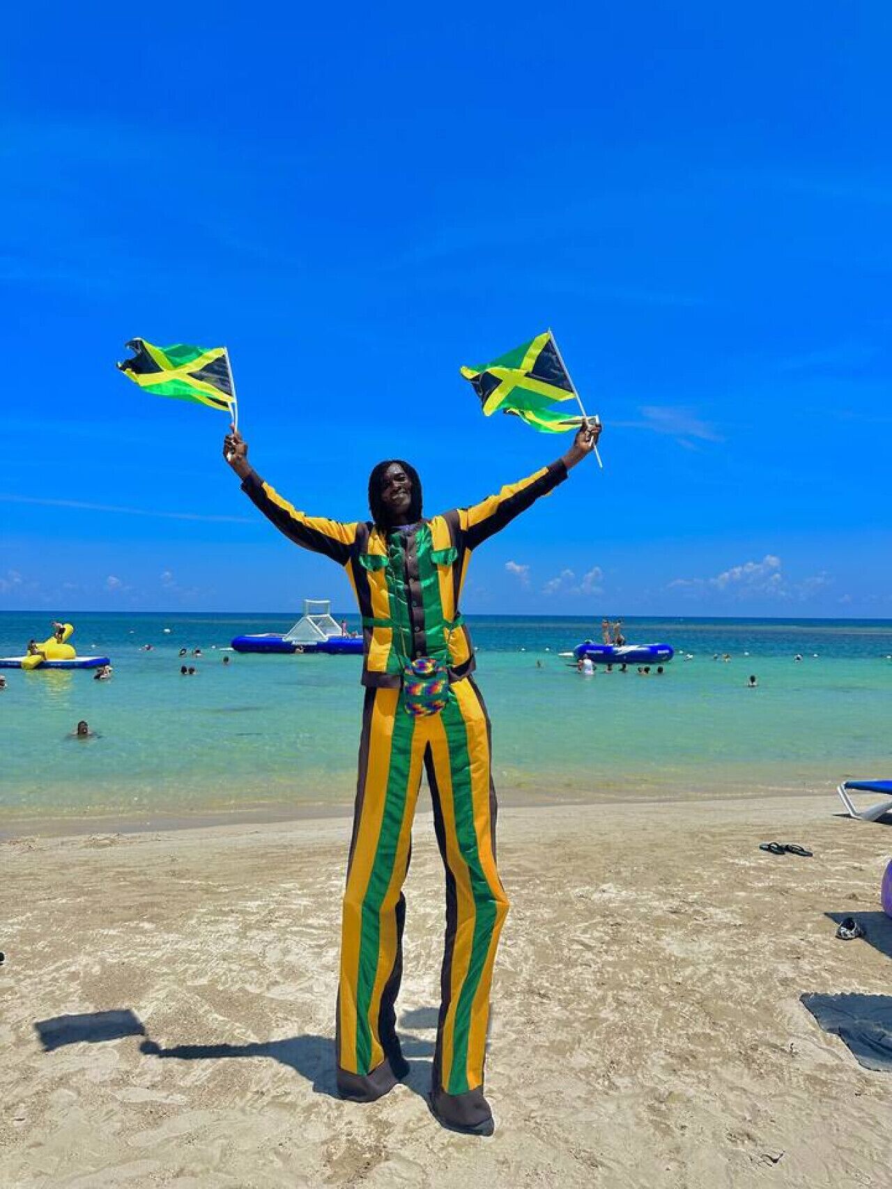 A person on a beach holding two flags, with clear blue skies and a sandy beach in the background.