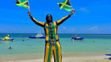 A person on a beach holding two flags, with clear blue skies and a sandy beach in the background.