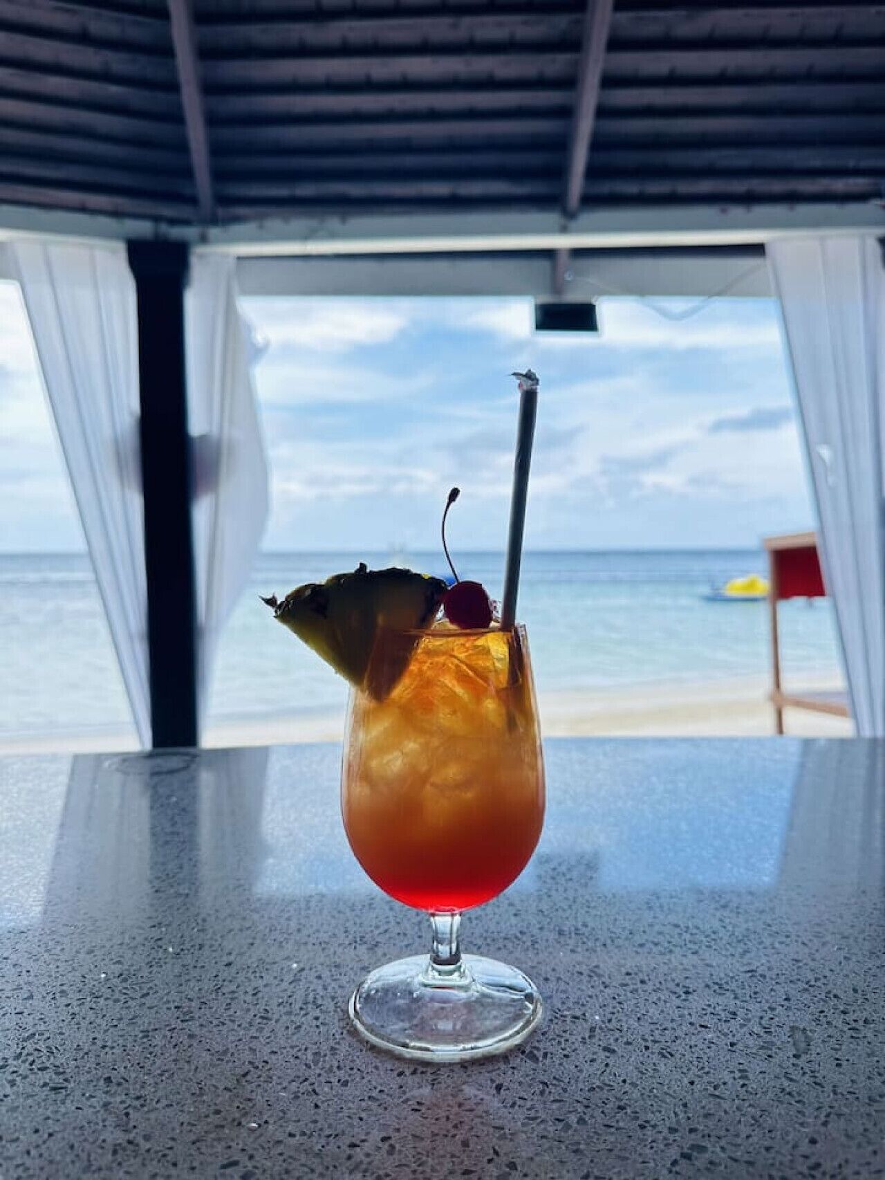 A cocktail glass with a cherry on a stick on a table by a beach.