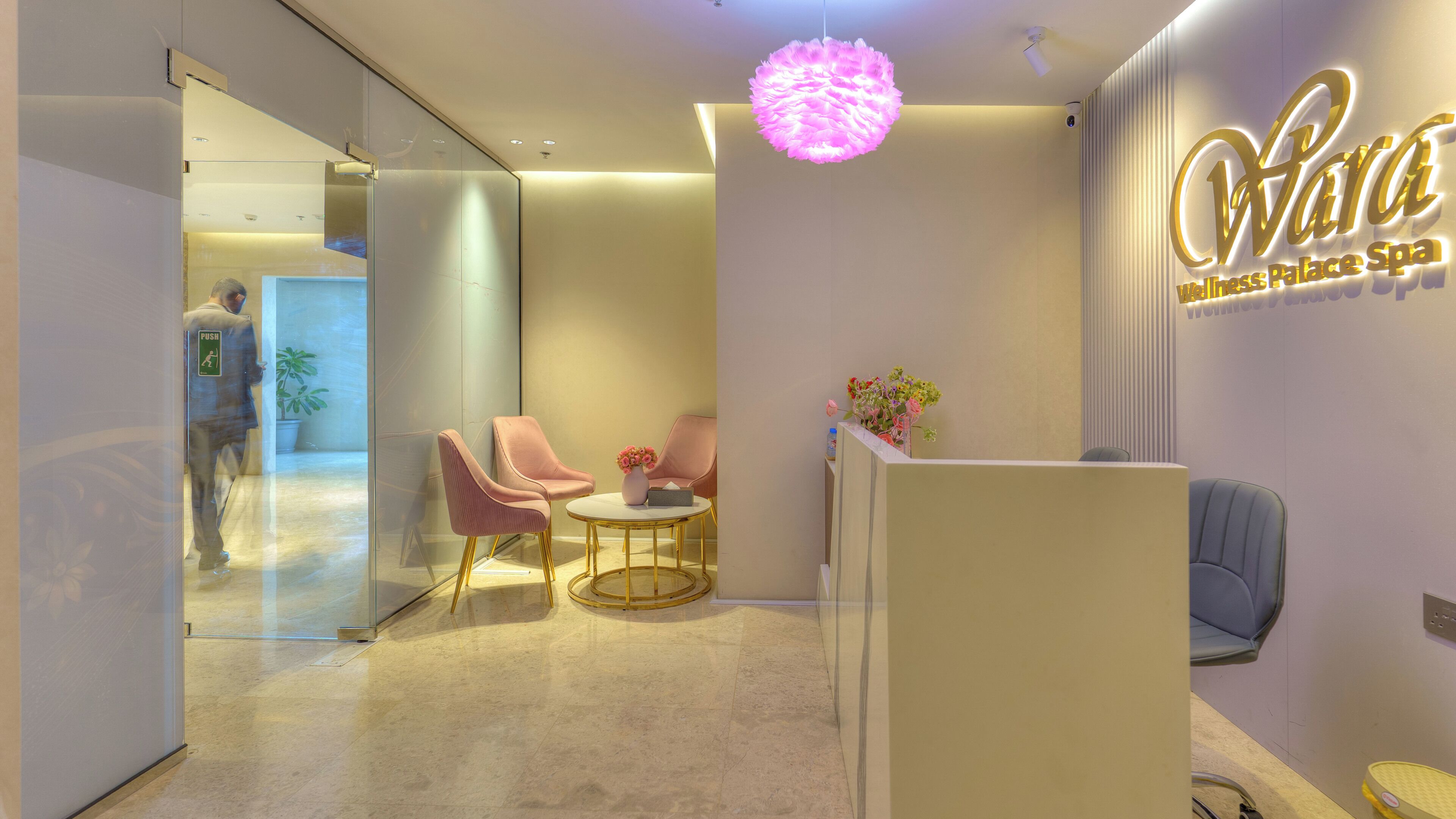 Zona de recepción con un letrero que dice 'Wara Wellness Palace Spa', dos sillones rosados, una mesa redonda y una planta en maceta.