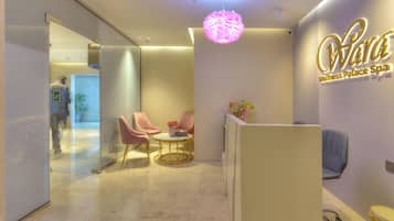 Zona de recepción con un letrero que dice 'Wara Wellness Palace Spa', dos sillones rosados, una mesa redonda y una planta en maceta.