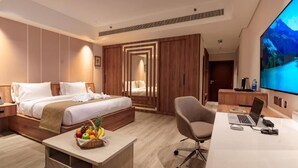 A modern hotel room with a bed, a desk with a laptop, a chair, a TV, and a fruit basket.