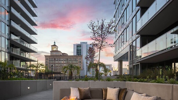 Eine Dachterrasse mit Feuerstelle mit Glasplatte, einem beigefarbenen Ecksofa und Blick auf ein historisches Gebäude im Hintergrund.