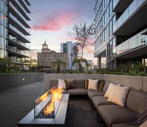 A rooftop lounge with a glass-top fire feature, a beige sectional sofa, and a view of a historic building in the background. - 35th-Floor Water Views: 2BR/2BA, Private Balcony & Pool (San Diego)