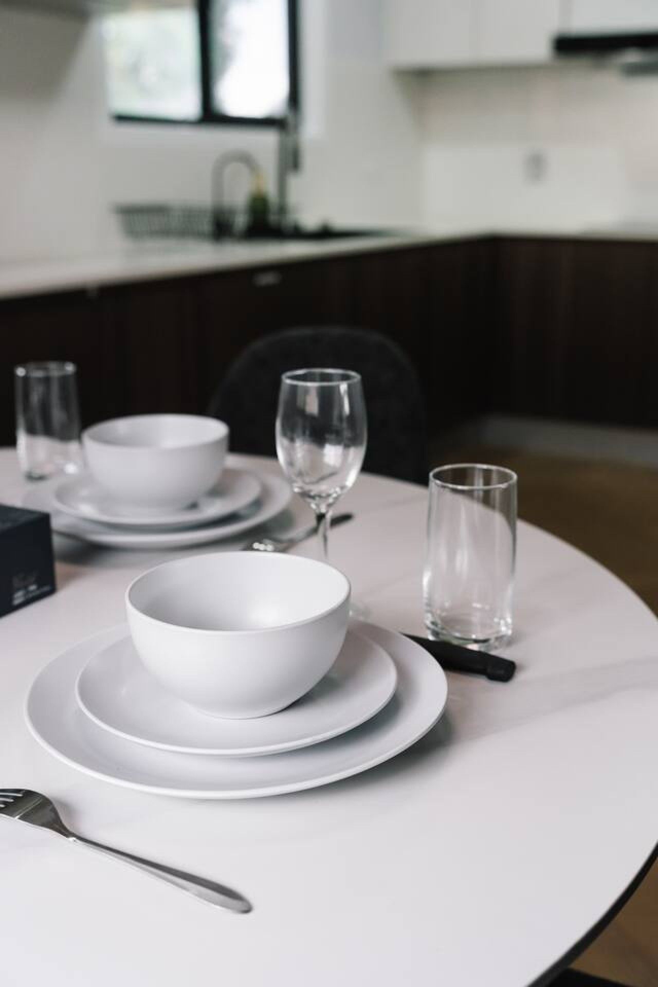 Une table à manger dressée avec de l’assiette blanche, des bols et des verres, avec une chaise et une cuillère visibles.