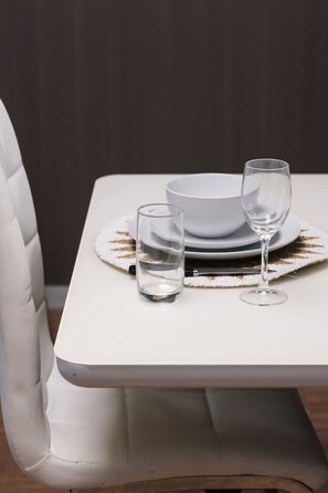Une table à manger avec un saladier blanc et deux verres, une chaise blanche et un mur foncé.