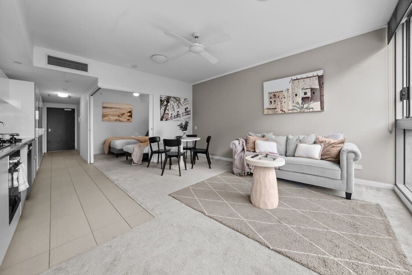 A modern living room with a grey sofa, a dining table with chairs, a kitchen area with cabinets, and a ceiling fan.