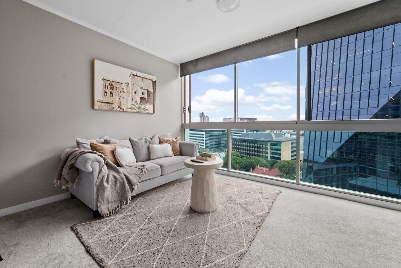 A modern living room with a grey sofa, a round coffee table, and a large window offering a city view.