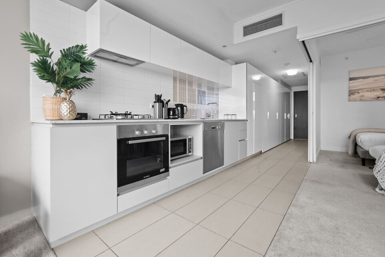 A modern kitchen with white cabinetry, a built-in oven, microwave, and dishwasher. There is a potted plant on the counter and a painting on the wall.
