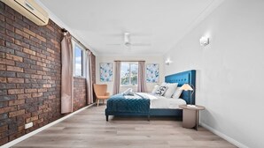 Een slaapkamer met een bakstenen muur, een bed met blauw hoofdeinde, een stoel en een nachtkastje met een lamp.