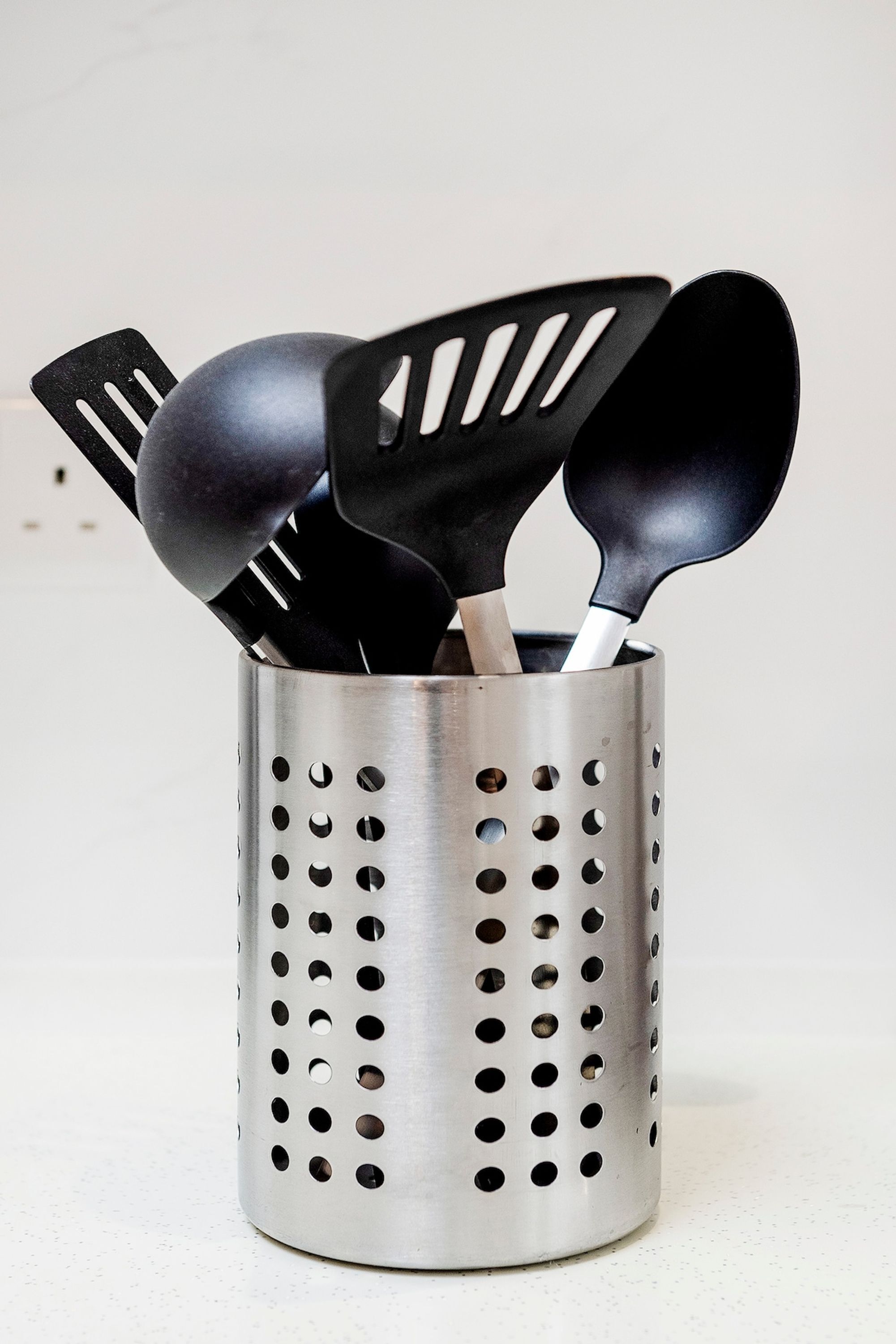 A stainless steel utensil holder with holes, containing black kitchen utensils.