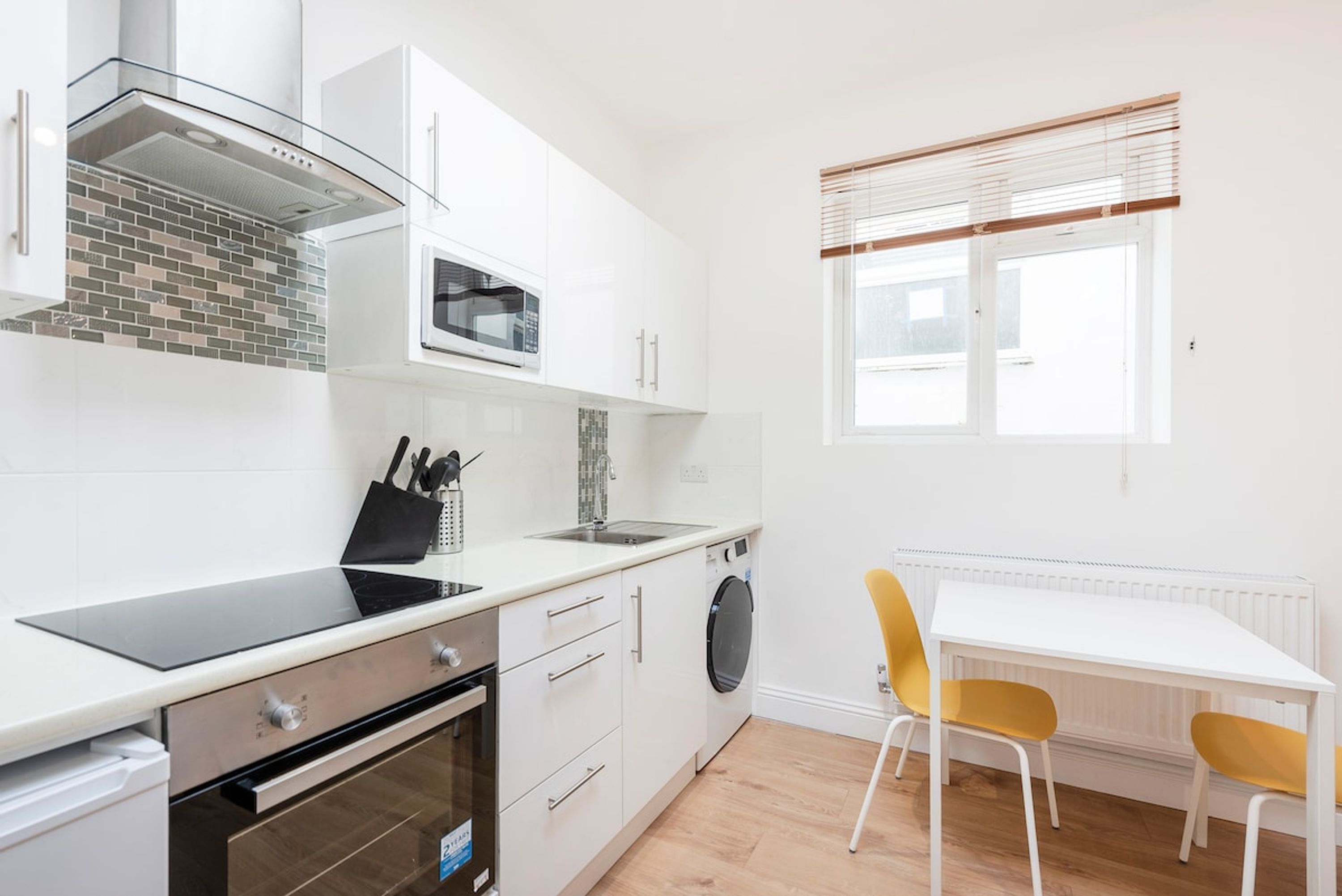 Eine kompakte Küche mit Mikrowelle, Backofen und Spüle, ein Essbereich mit Tisch und Stühlen sowie ein Fenster mit Jalousien.