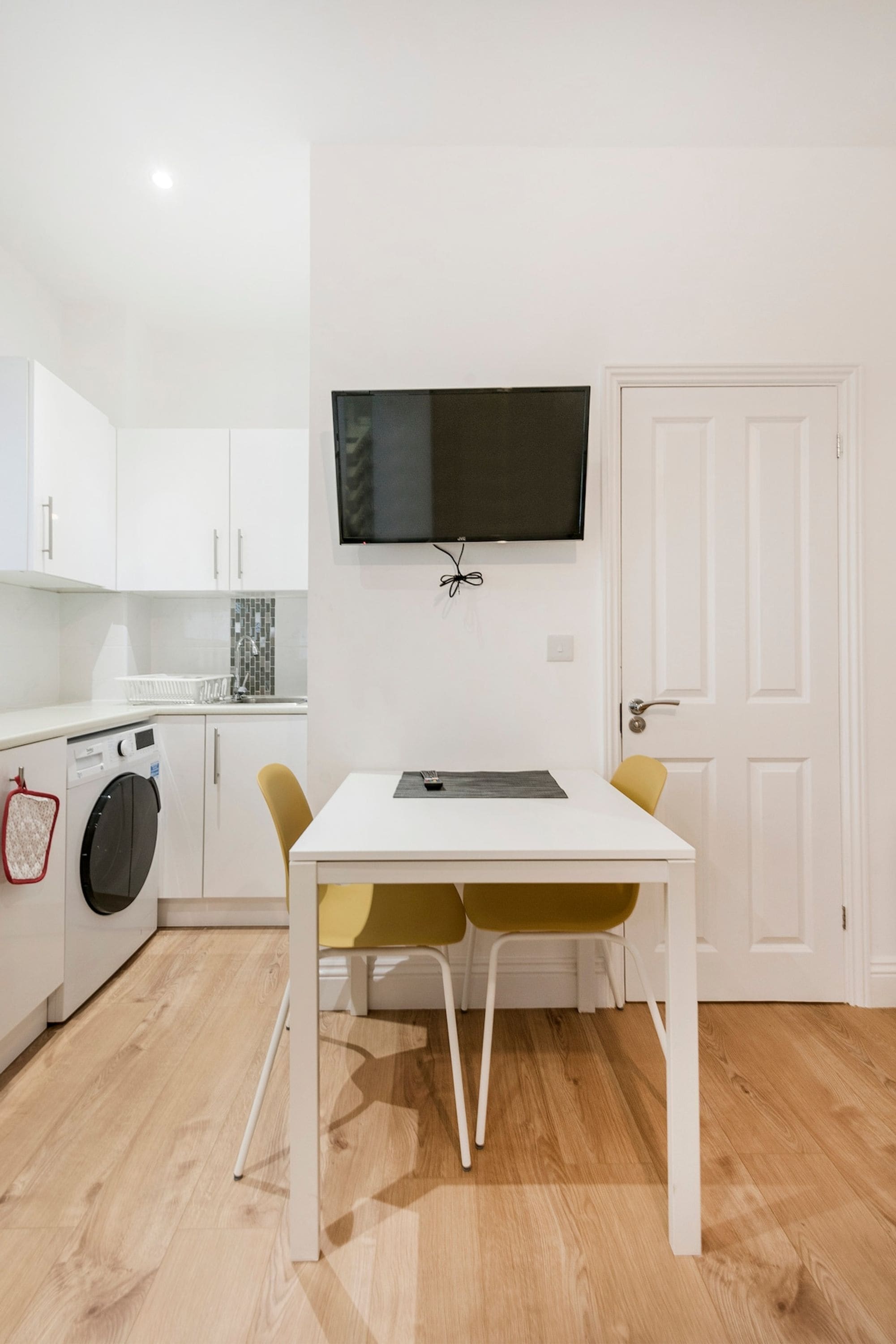 A compact kitchen with a washing machine, a dining table with chairs, a flat-screen TV mounted on the wall, and a white door.