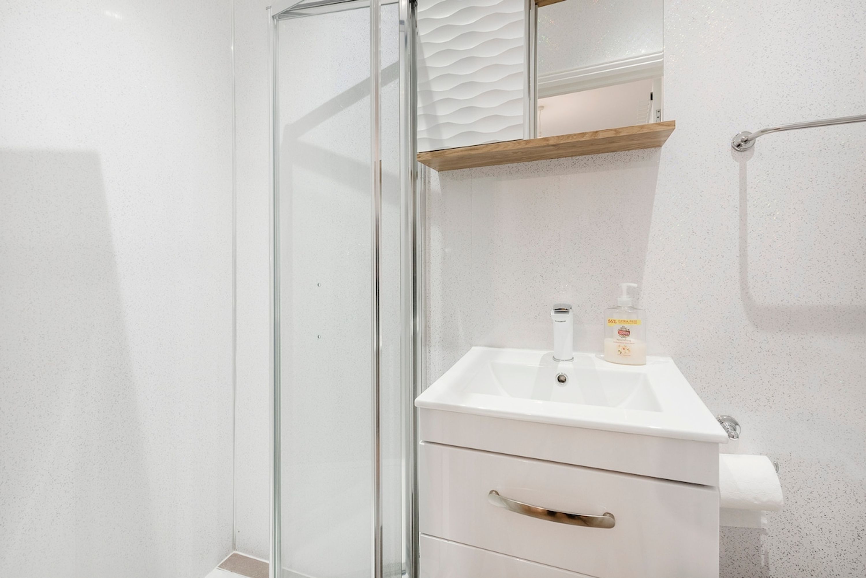 A bathroom with a glass shower enclosure, a white sink with a soap dispenser, and a wooden shelf above the sink.