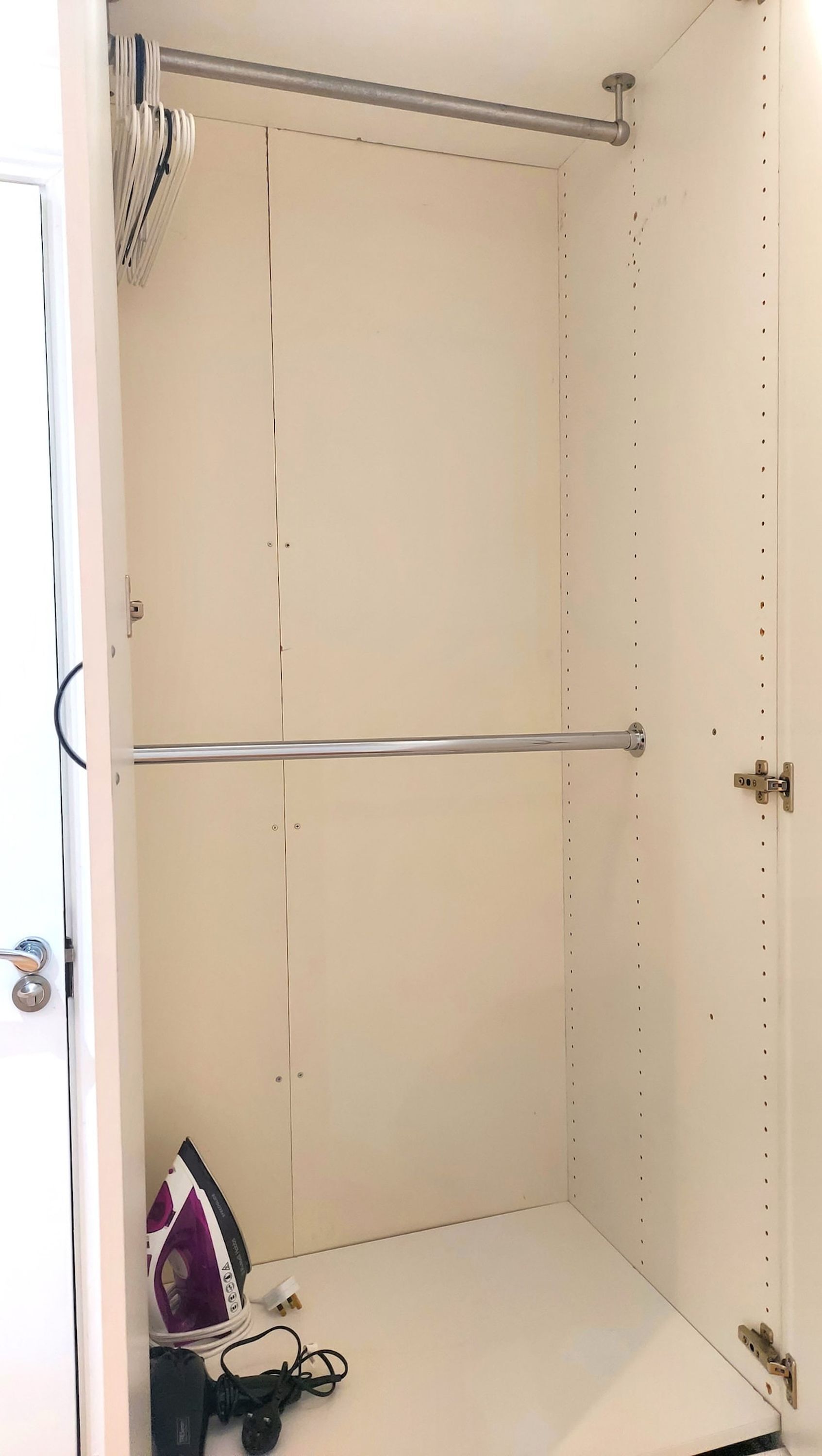 A white wardrobe with a metal rod across the top, a door with a lock, and a small ironing board with an iron and a power strip on the floor.