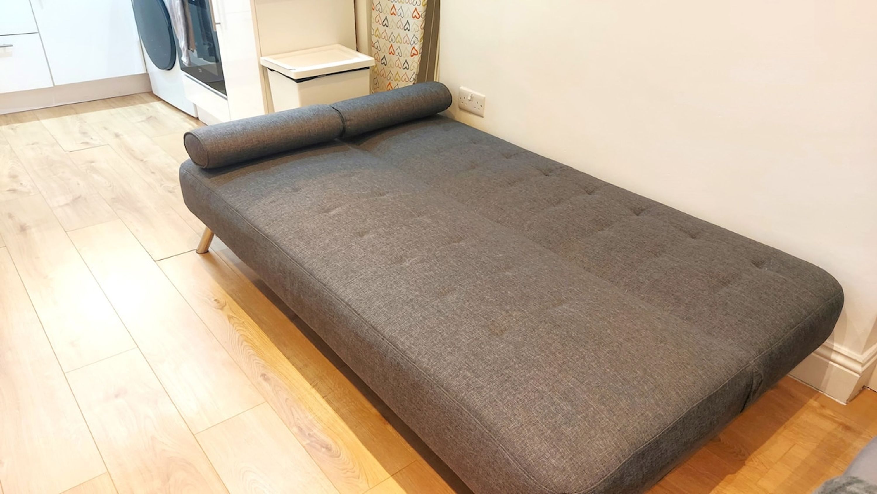 Una habitación pequeña con cama individual, colchón gris, suelo de madera, lavadora y una mesita de noche.