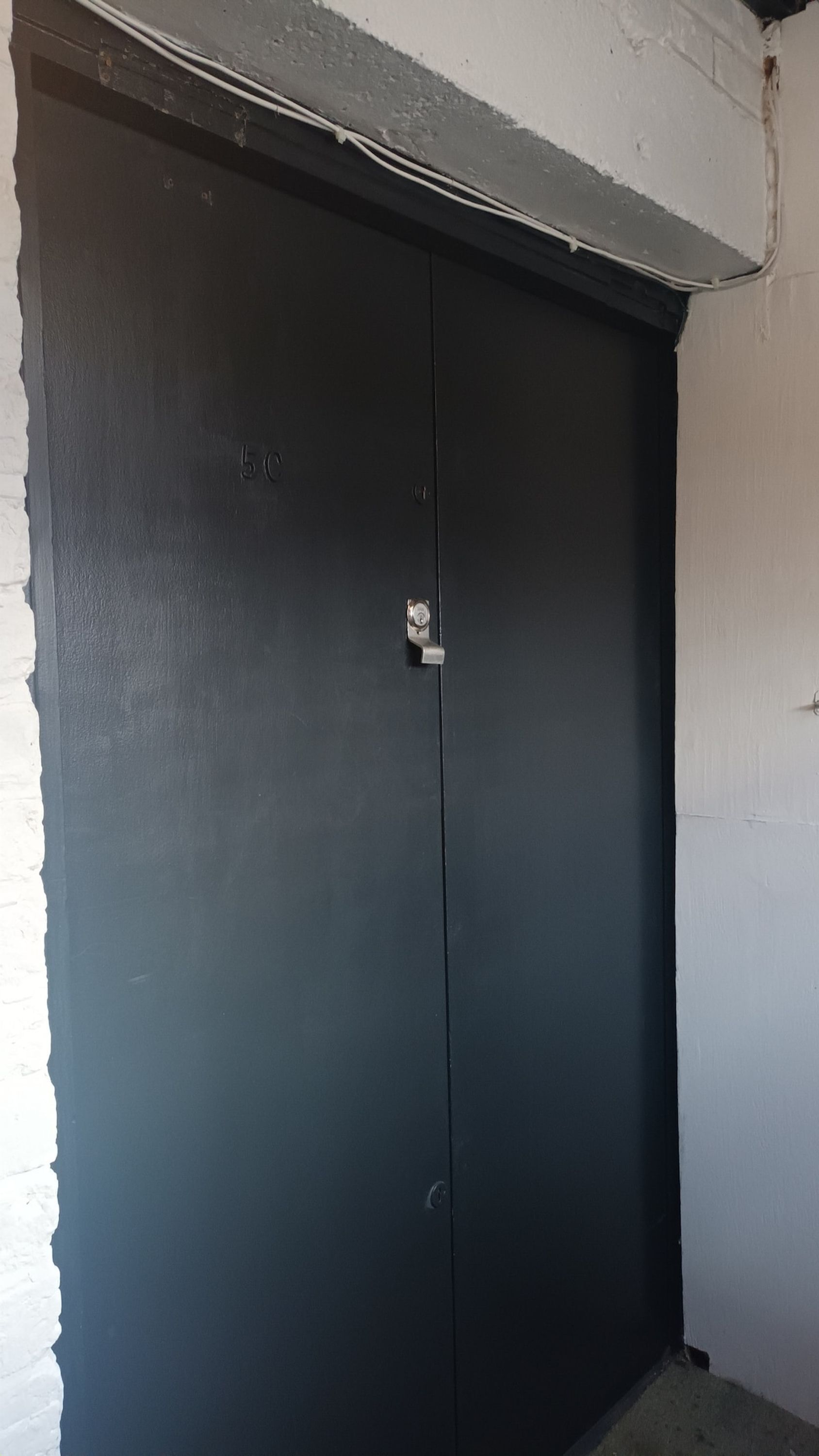 Une porte noire fermée, dotée d’une petite serrure et d’un trou de serrure, située dans un coin, avec un mur blanc et un sol en béton.