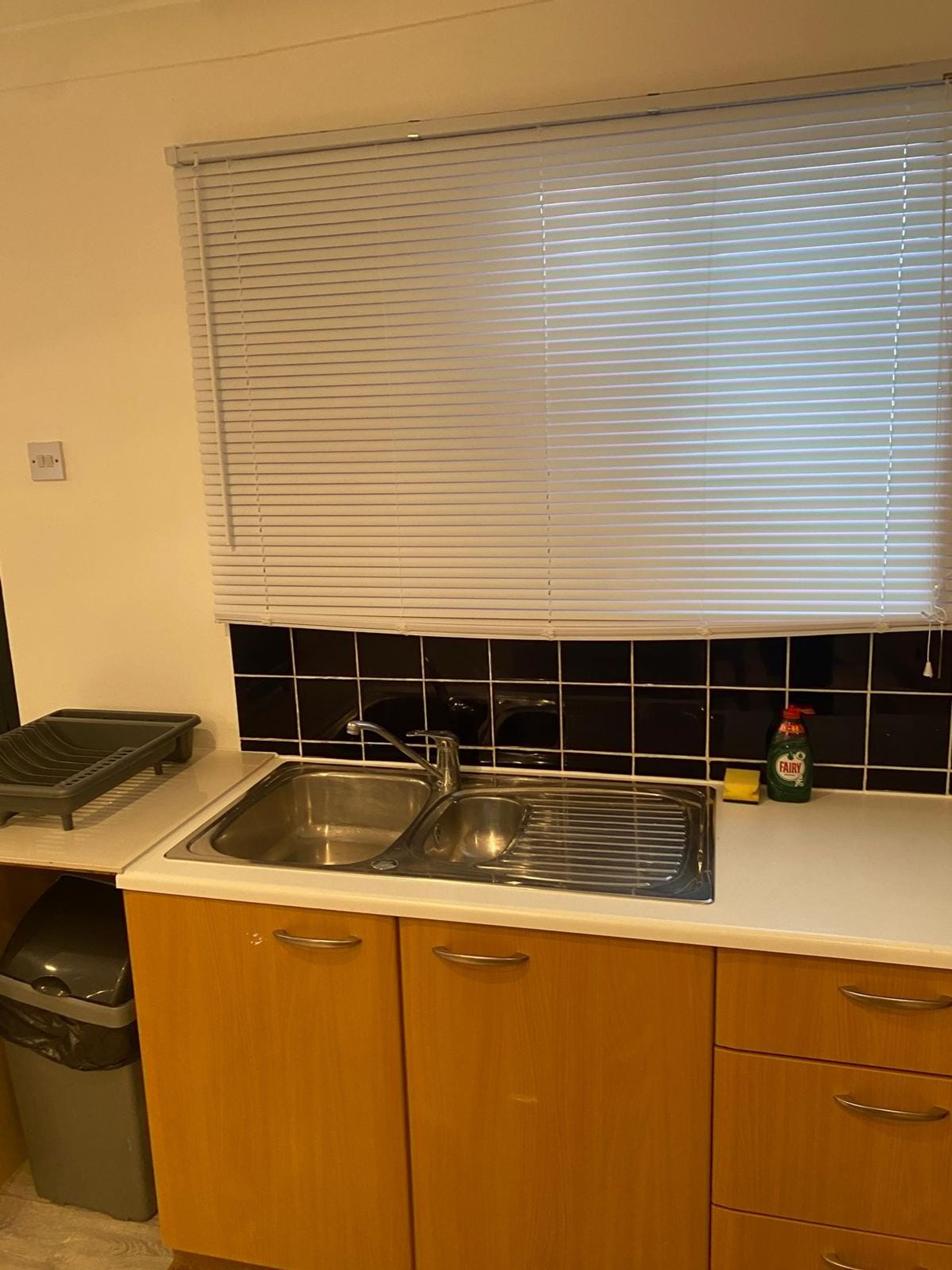 A kitchen with a sink, a dish rack, a trash bin, and a window with blinds.