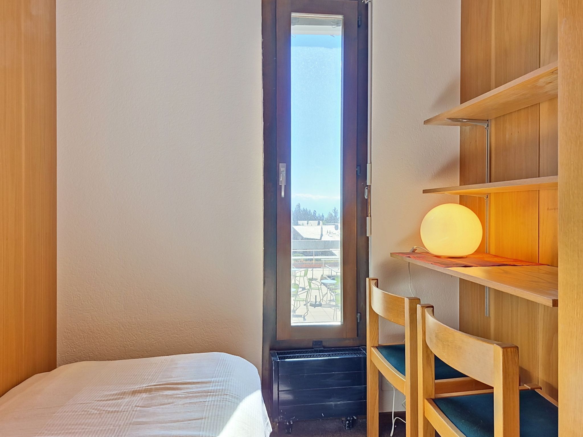 Een kamer met een bed, een houten stoel, een plank met een lamp en een raam met uitzicht.