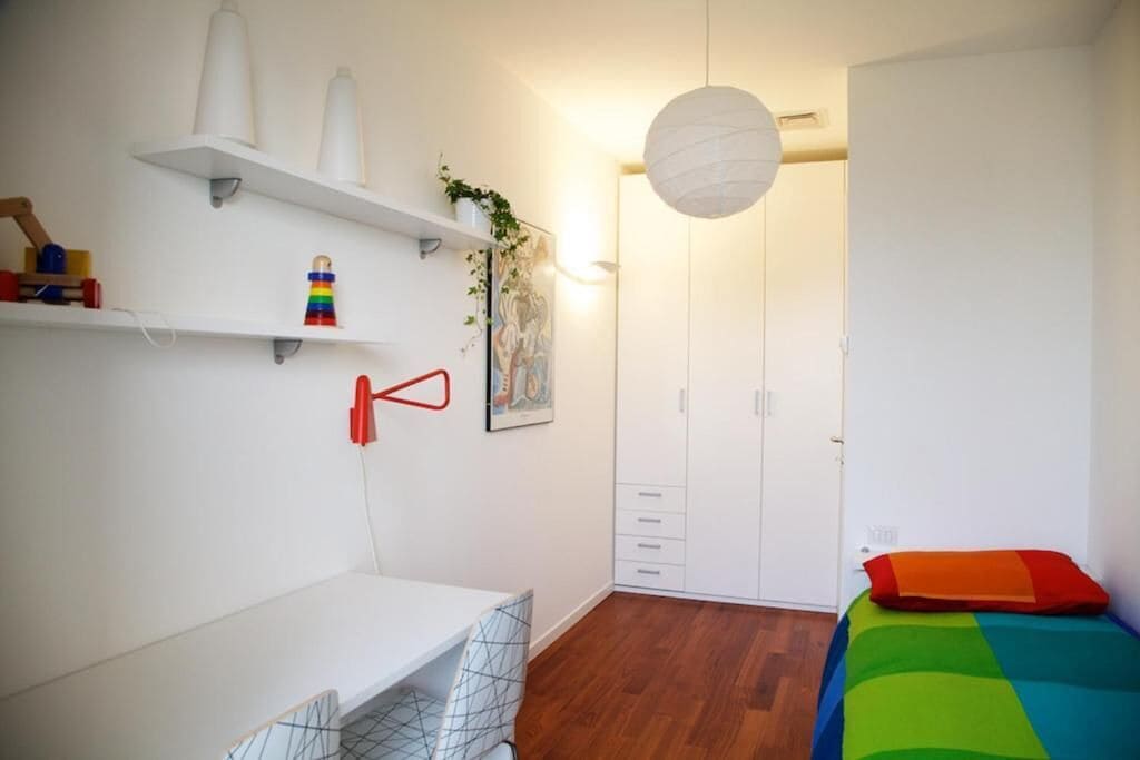 Ein Schlafzimmer mit Bett, Schreibtisch, Kleiderschrank und Regalen mit Gegenständen.