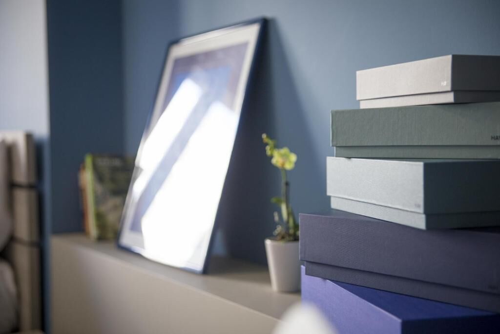 Une pile de livres sur une étagère, avec un tableau encadré appuyé contre le mur.