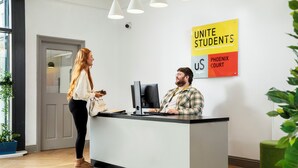 A reception desk with a computer, a sign reading 'UNITE STUDENTS US PHOENIX COURT', and a door in the background. - Student Only Phoenix Court (Bristol)