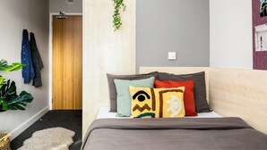 A bedroom with a bed, pillows, a plant, a door, and a wall with a shelf. - Student Only Phoenix Court (Bristol)
