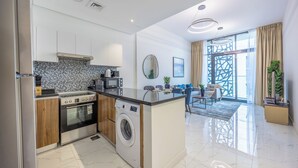 A modern kitchen with a built-in washing machine, a dining area with a glass table, and a living room with a sofa and a large window. - Bloomfields 2BR in The Gate - Masdar  (Abu Dhabi)