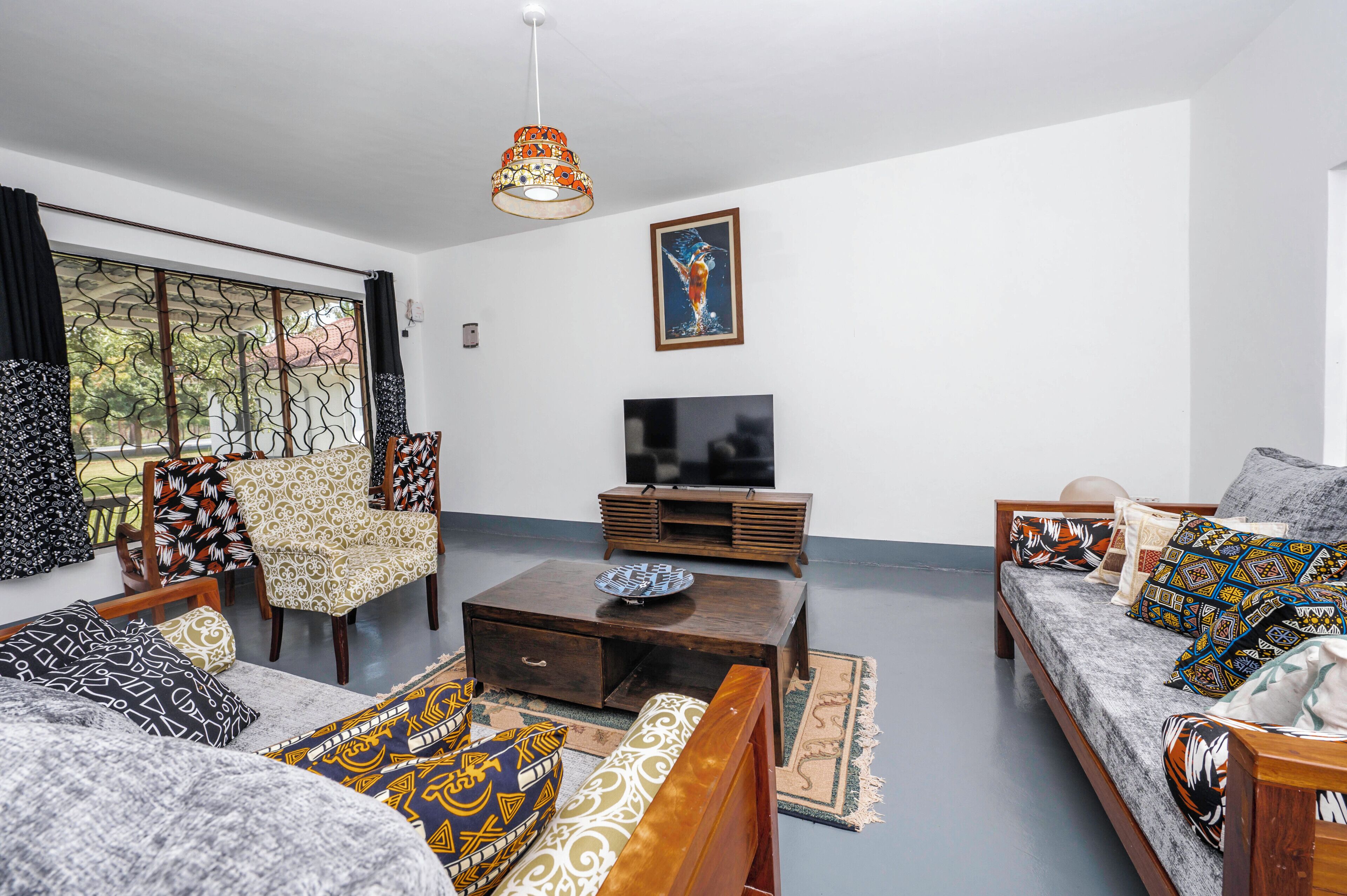Ein Wohnzimmer mit einer Couch, Sesseln, einem Couchtisch, einem Fernseher und einem großen Fenster mit Vorhängen.