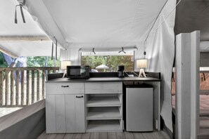 Una kitchenette compacta con microondas, cafetera y lámpara, bajo un techo blanco cubierto, con vistas a la vegetación.