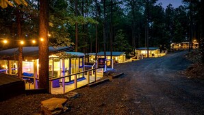 Un hôtel de style rustique, aménagé comme un chalet, comprenant plusieurs chambres, un espace extérieur couvert avec des sièges et un sentier de terre qui traverse la forêt.