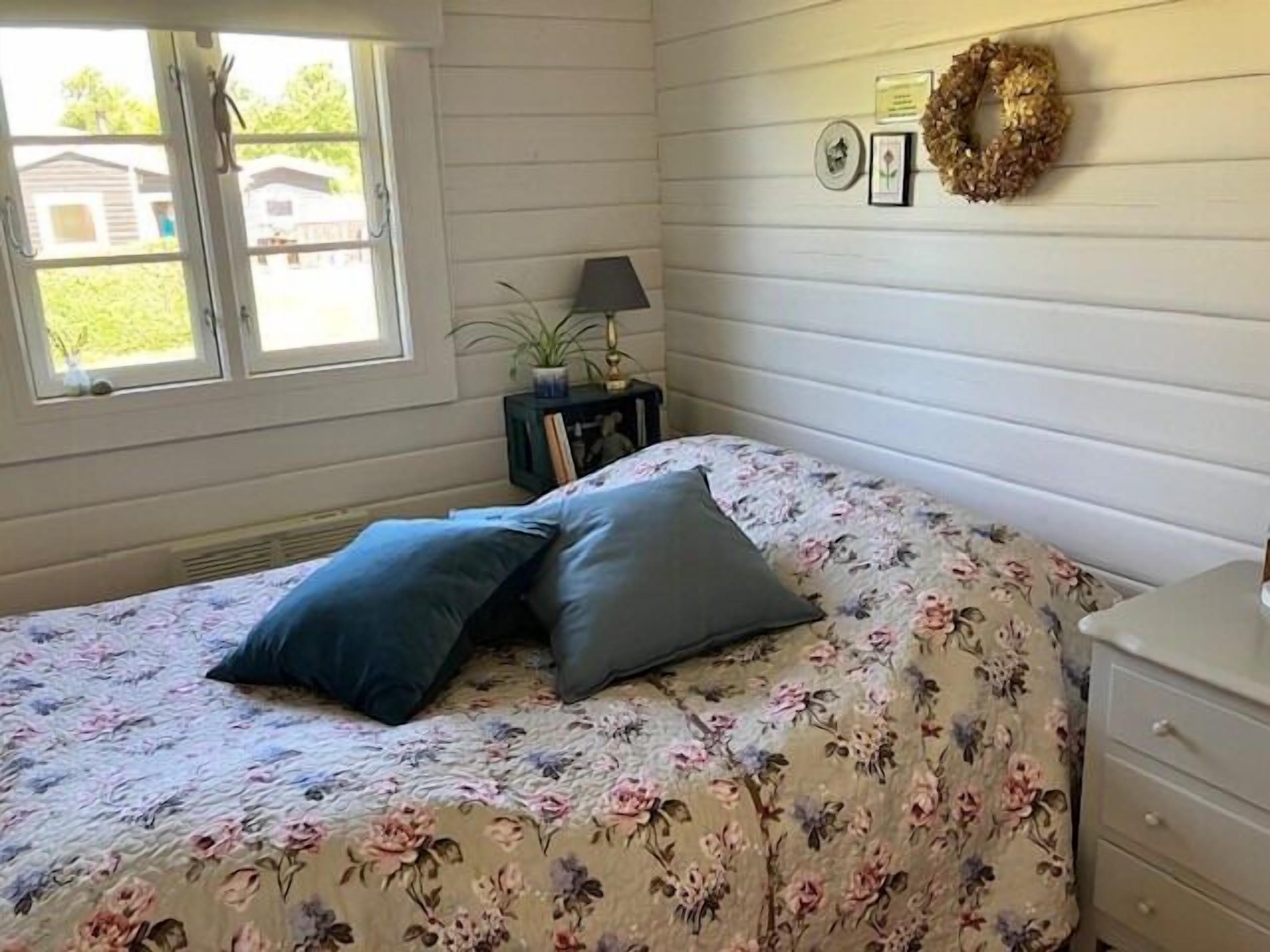 Een slaapkamer met een bloemenbedekking, twee kussens, een nachtkastje met een lamp, een kleine plant en een aan de muur bevestigd krans.
