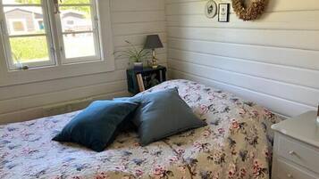 Een slaapkamer met een bloemenbedekking, twee kussens, een nachtkastje met een lamp, een kleine plant en een aan de muur bevestigd krans.