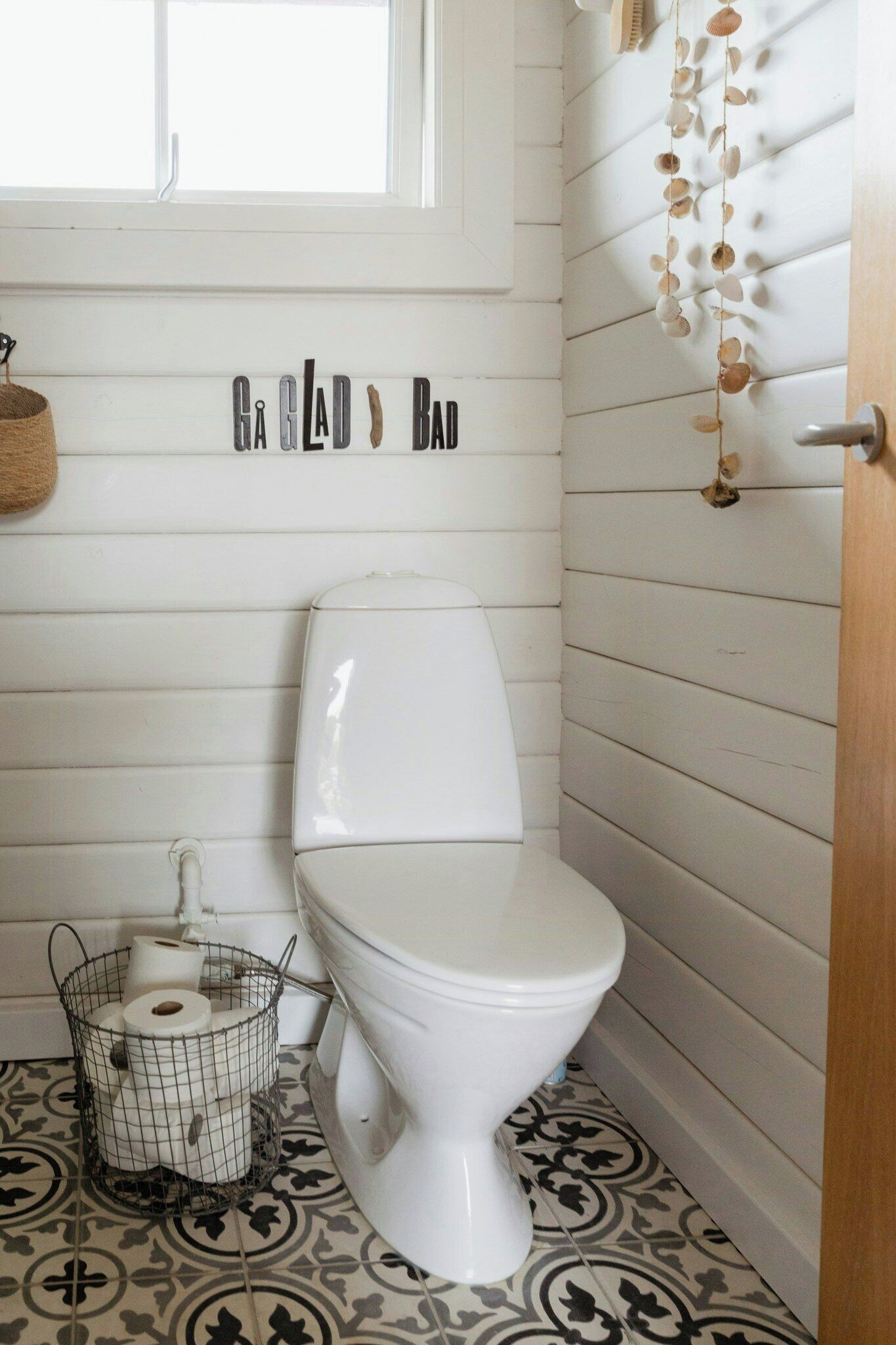 A bathroom with a white toilet, patterned floor tiles, a hanging decoration, and a wall-mounted sign.