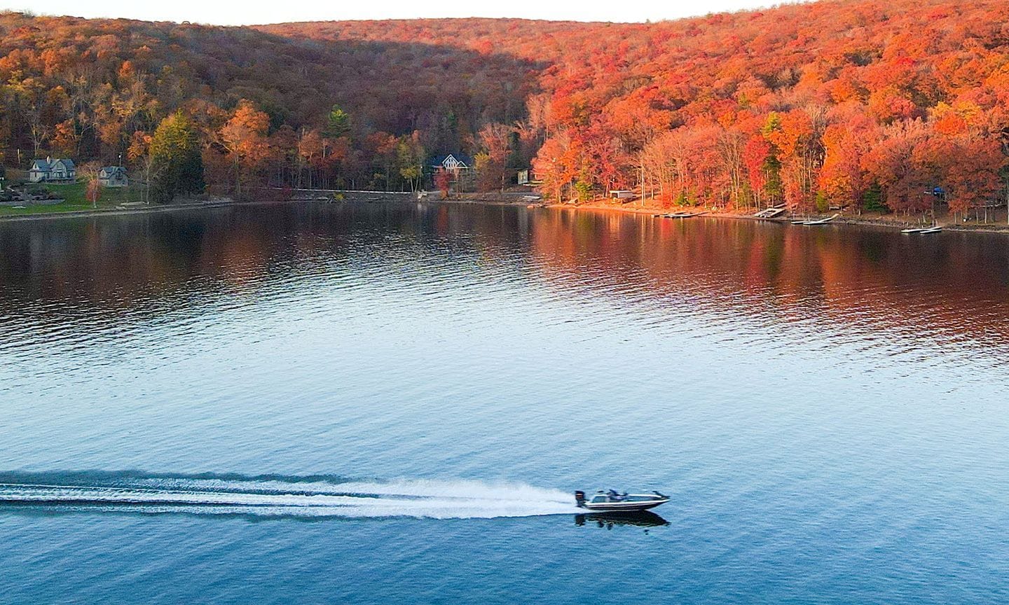 A lake with a boat, surrounded by autumn-colored trees and houses in the background.