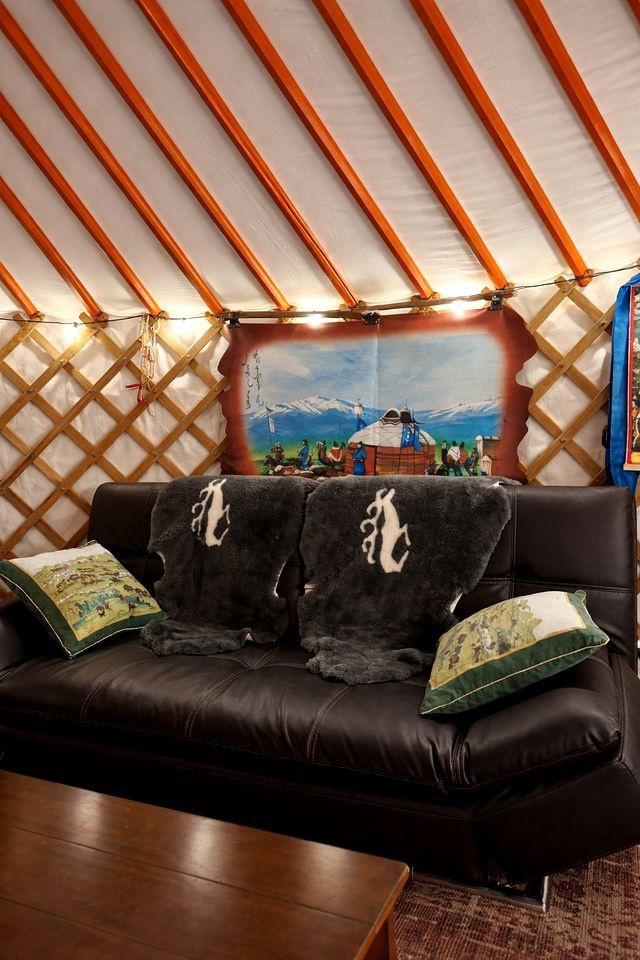 A room with a black leather sofa, two pillows, a wooden table, and a large window with a view of mountains and a traditional yurt.