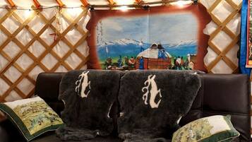 A room with a black leather sofa, two pillows, a wooden table, and a large window with a view of mountains and a traditional yurt.