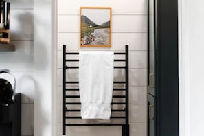 A towel rack with a white towel, a framed landscape photo, and a black towel rack in a modern bathroom. - Cute Elevated Tiny House with Private Hot Tub for Romantic Escapes in Alabama (Guntersville)