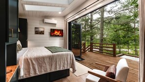 Un dormitorio con cama, televisión y chimenea. Cuenta con un balcón con cubierta de madera y vista a árboles.
