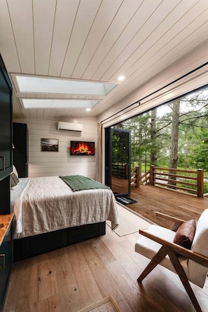 A bedroom with a bed, a TV, a fireplace, and a balcony with a view of trees.