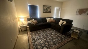 Una sala de estar con un sofá modular, un tapete estampado, una mesita de noche con una lámpara y un cuadro en la pared.