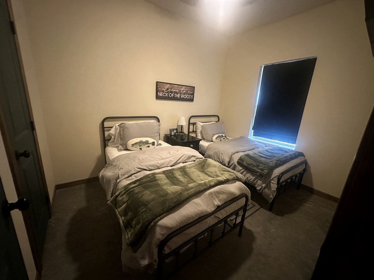 Quarto com duas camas, mesa de cabeceira com luminária, janela com persianas e um letreiro de boas-vindas na parede.