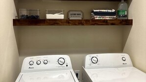 A laundry room with two white front-loading washing machines, a shelf with folded towels, a box of laundry detergent, and a sign indicating the room's purpose.