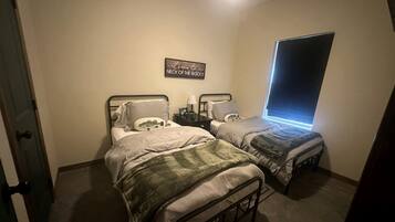 A bedroom with two beds, a bedside table with a lamp, a window with blinds, and a welcome sign on the wall.